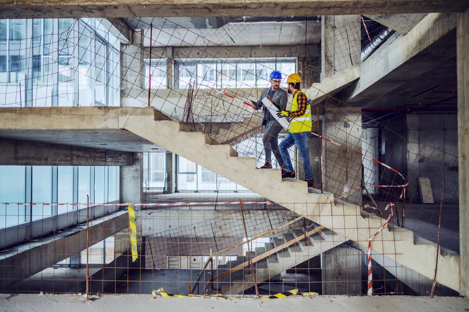 Two construction workers in protective gear discuss plans on stairs in an unfinished building, surrounded by safety nets and barriers.