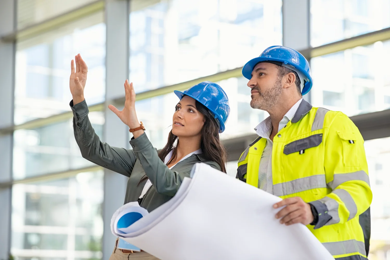Two construction workers in hard hats review blueprints and discuss details inside a modern building.