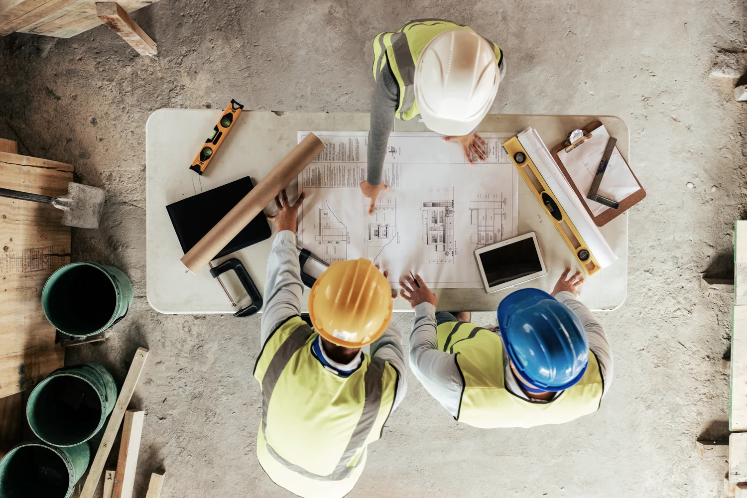 Three construction workers in helmets and vests review blueprints on a table. Various tools and devices are scattered around. Top-down view.