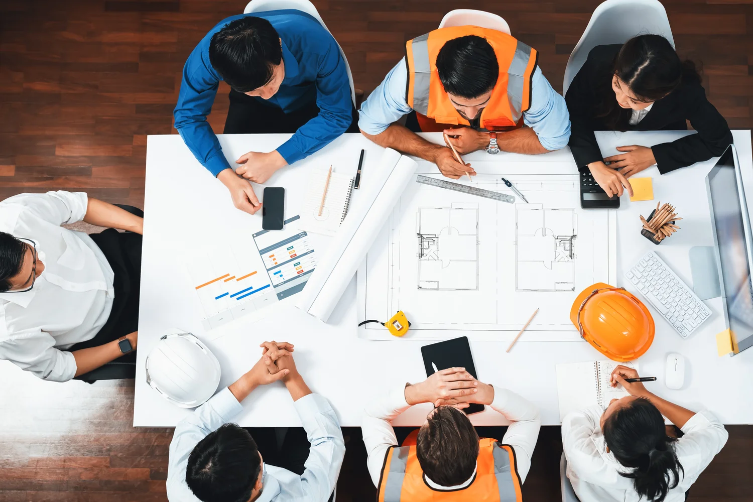 Group of people in a meeting around a table with construction plans, digital tablets, and hard hats. Some wear business attire, others in safety vests.