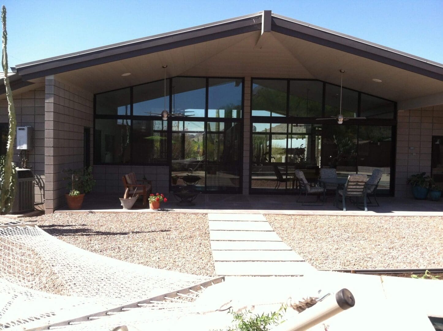 Modern house with large glass windows and sloped roof, featuring a patio with seating and a hammock in the foreground. Desert landscaping with gravel and cacti.