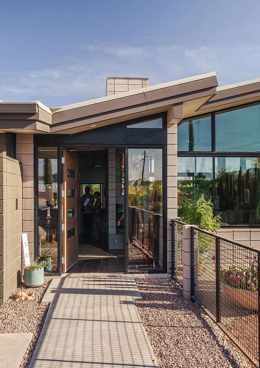 cr=t_0% (5) Modern house entrance with angular roof, glass windows, and concrete block walls. A pathway leads to a glass and wood door. There are plants on the sides and a fence along the right side.