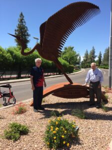 Two elderly men stand beside a large eagle sculpture outdoors.